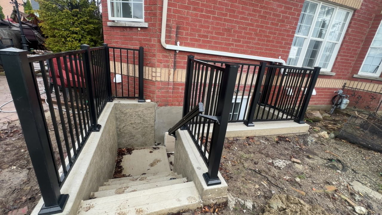 Wide view of a poured concrete basement walkout with landing, stairs and railings against a red brick house